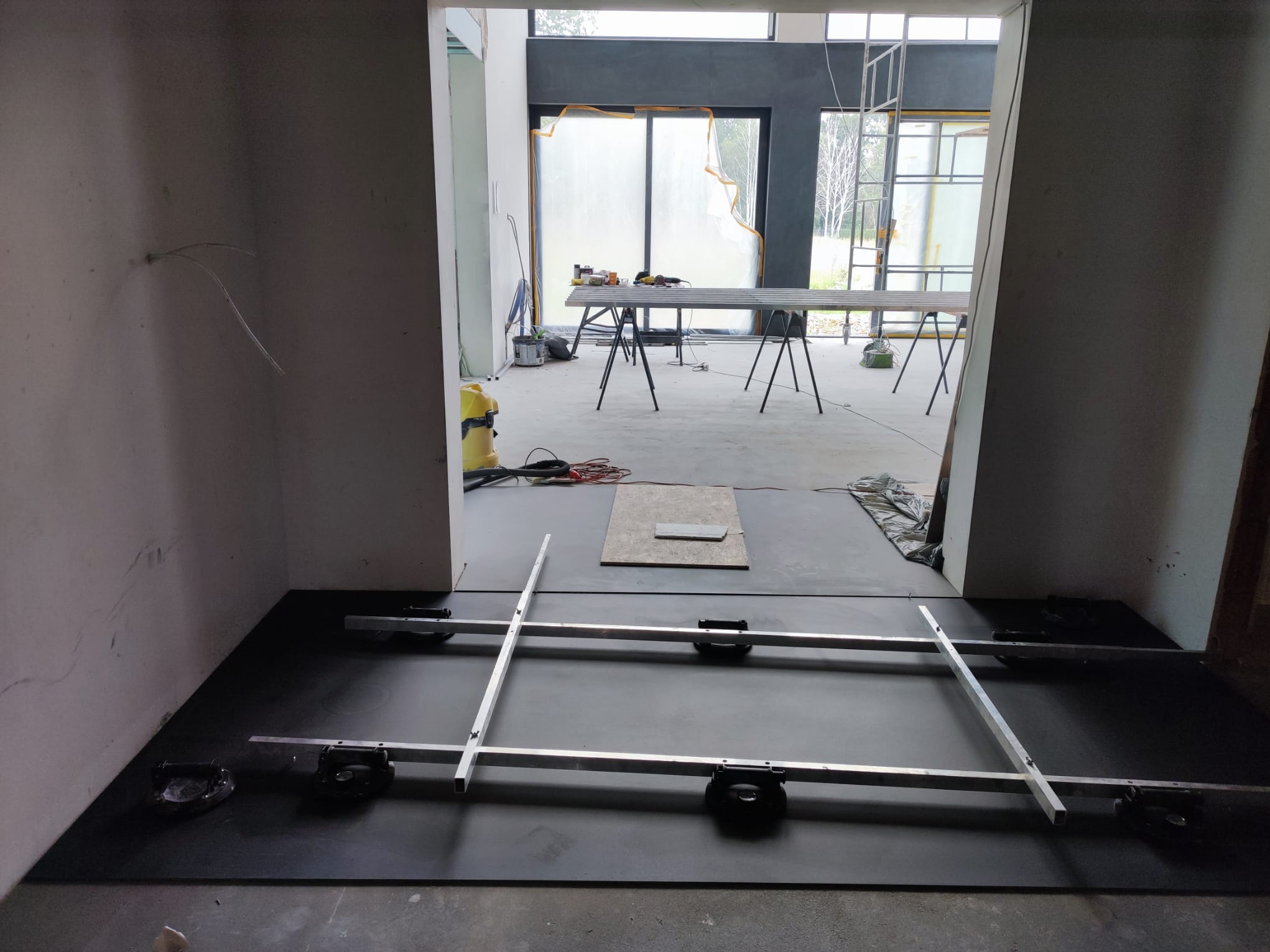 Przygotowanie podłoża: czarna mata wygłuszająca z aluminiową konstrukcją poziomującą, narzędzia w tle, surowe ściany i okna zabezpieczone folią. Wnętrze w trakcie prac.