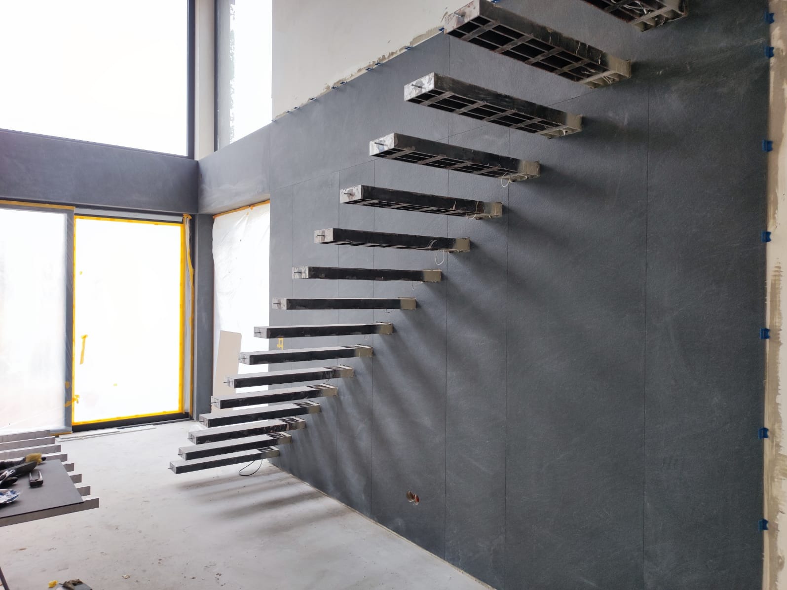 Montaż schodów wspornikowych na ścianie wyłożonej szarymi płytami. Widoczne elementy mocujące i surowe wykończenie wnętrza w jasnym pomieszczeniu.