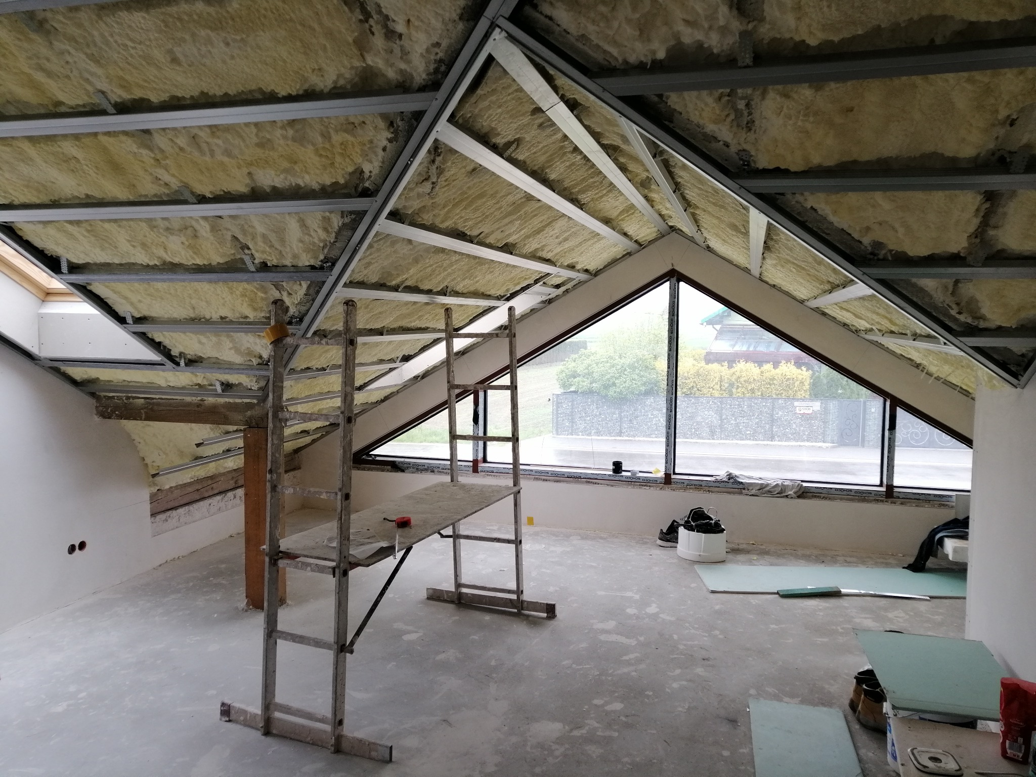 Poddasze w trakcie izolacji: wełna mineralna między metalowymi profilami sufitu, rusztowanie, okno z widokiem na zewnątrz. Etap prac wykończeniowych.