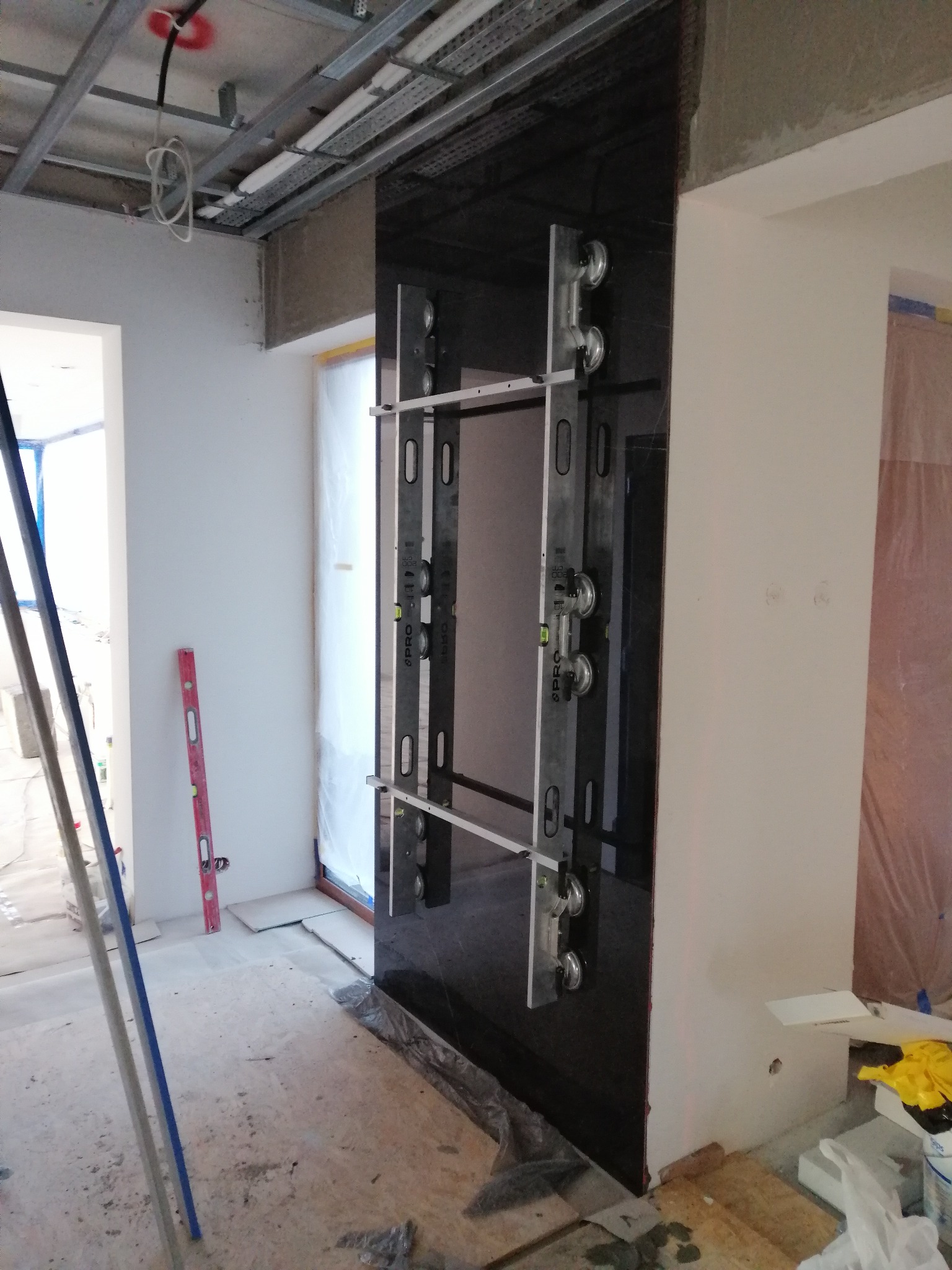 Montaż czarnej, błyszczącej płyty wielkoformatowej na ścianie, przy użyciu przyssawek i poziomicy. Widoczne narzędzia glazurnicze i surowy stan wykończenia wnętrza.