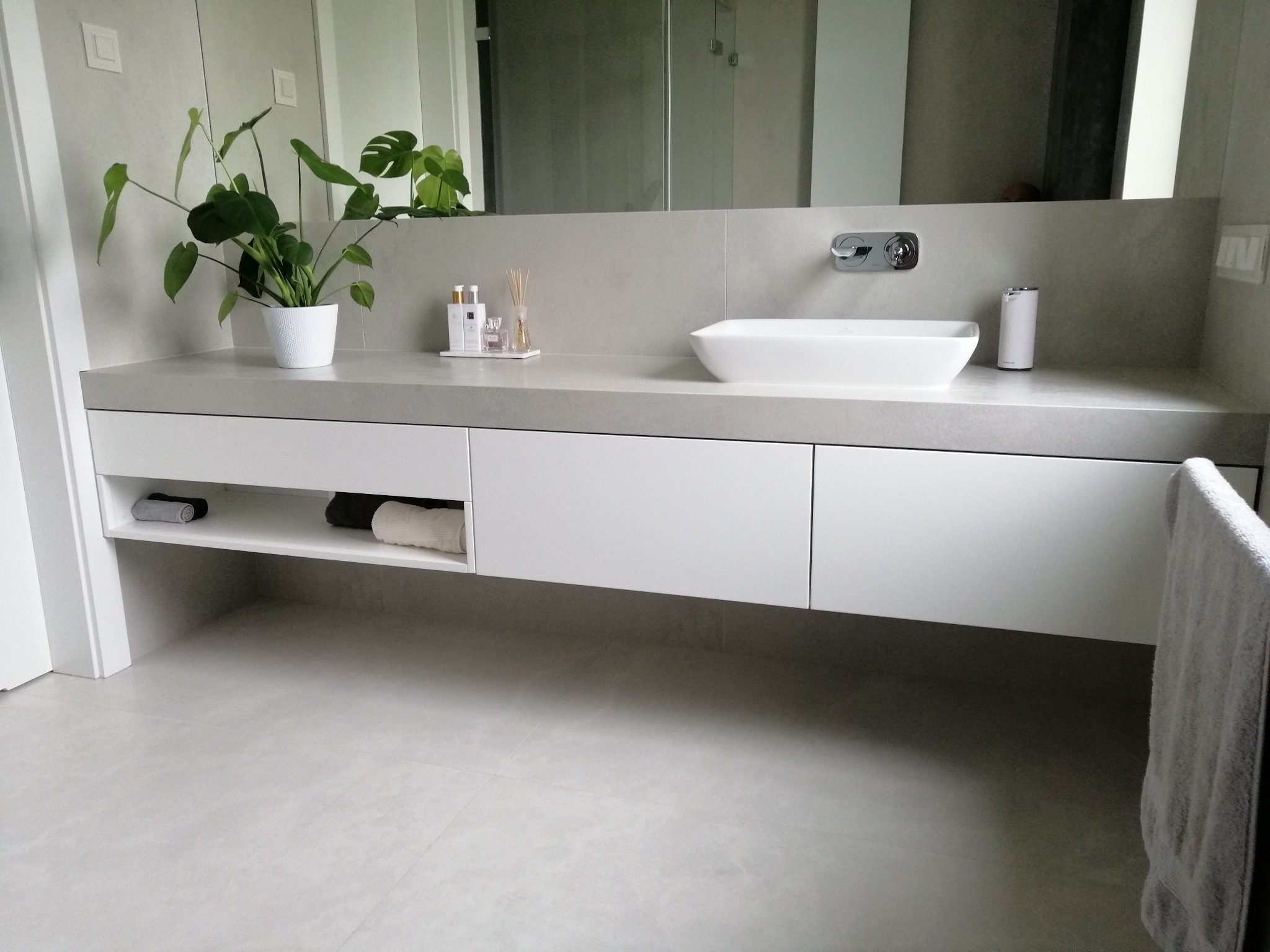 Nowoczesna łazienka z szarymi płytkami, białą umywalką nablatową i szafką pod umywalką. Na blacie dekoracje i roślina w doniczce. Minimalistyczny styl.