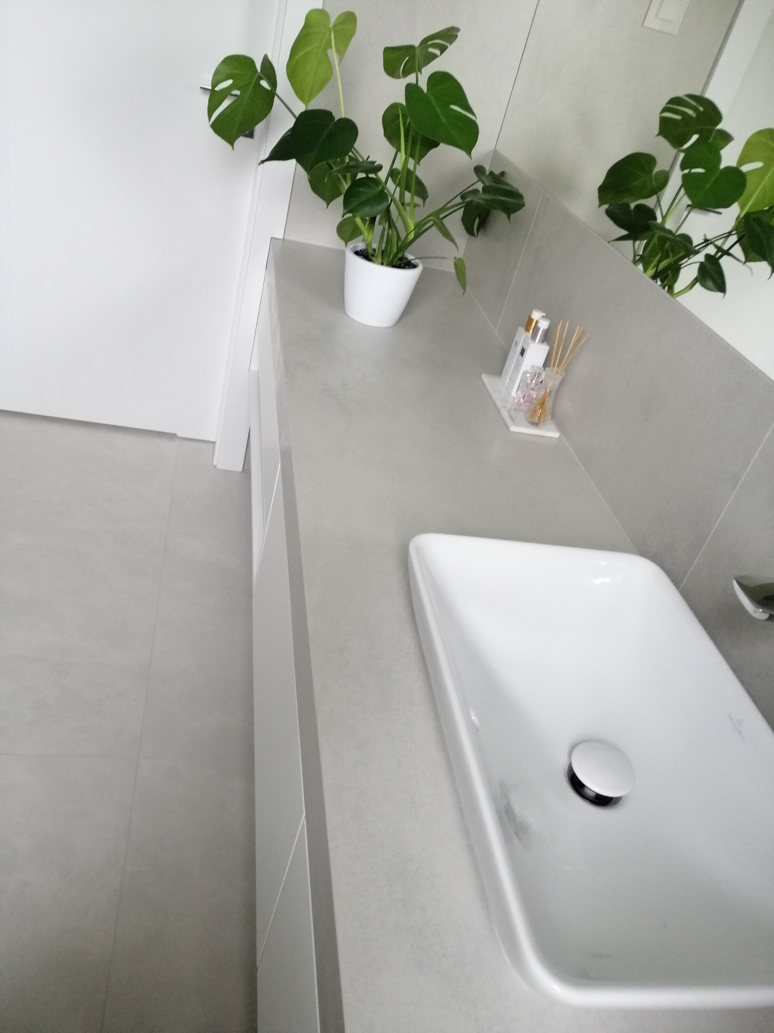 Minimalistyczna łazienka z białą umywalką nablatową, szarą płytką i zieloną rośliną monstera. Oświetlenie górne. Widoczne detale wykończenia.