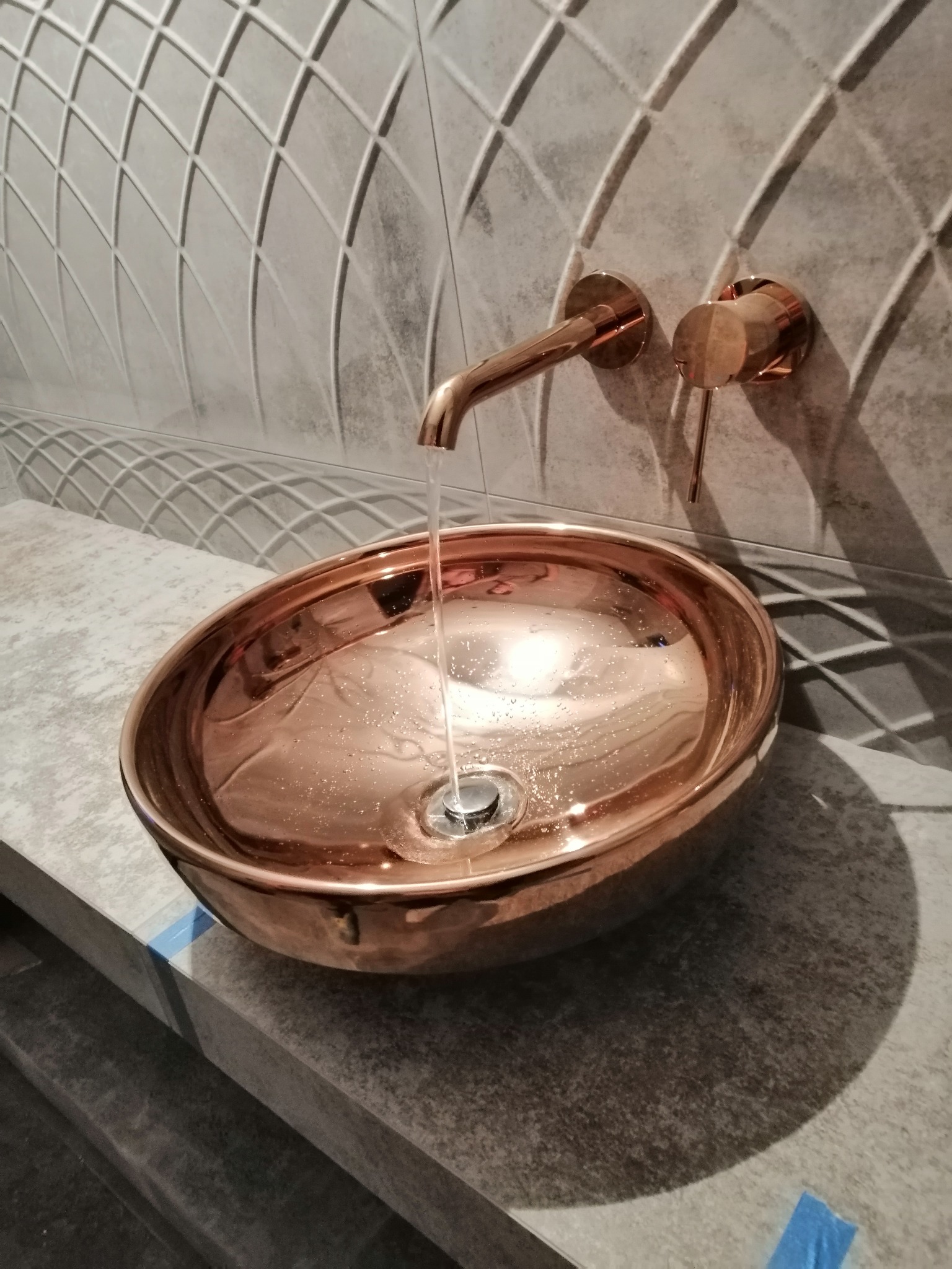 Miedziana umywalka nablatowa z armaturą w kolorze różowego złota na tle szarej ściany z geometrycznym wzorem. Woda leje się z kranu.
