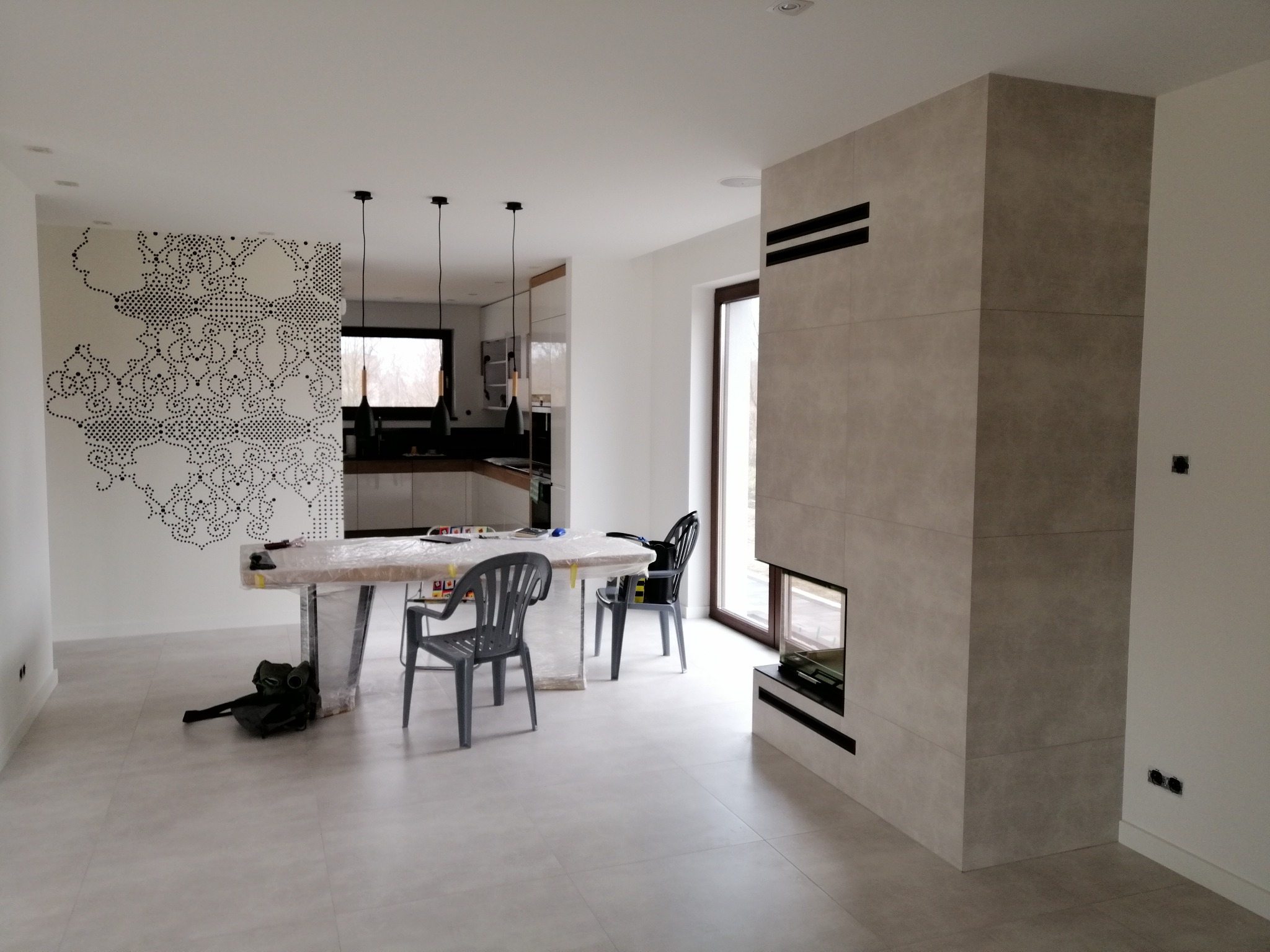 Jasne płytki podłogowe w salonie z kominkiem obłożonym płytkami imitującymi kamień. Na ścianie dekoracja w postaci wzoru z kropek. Widoczne krzesła i stół przykryty folią.