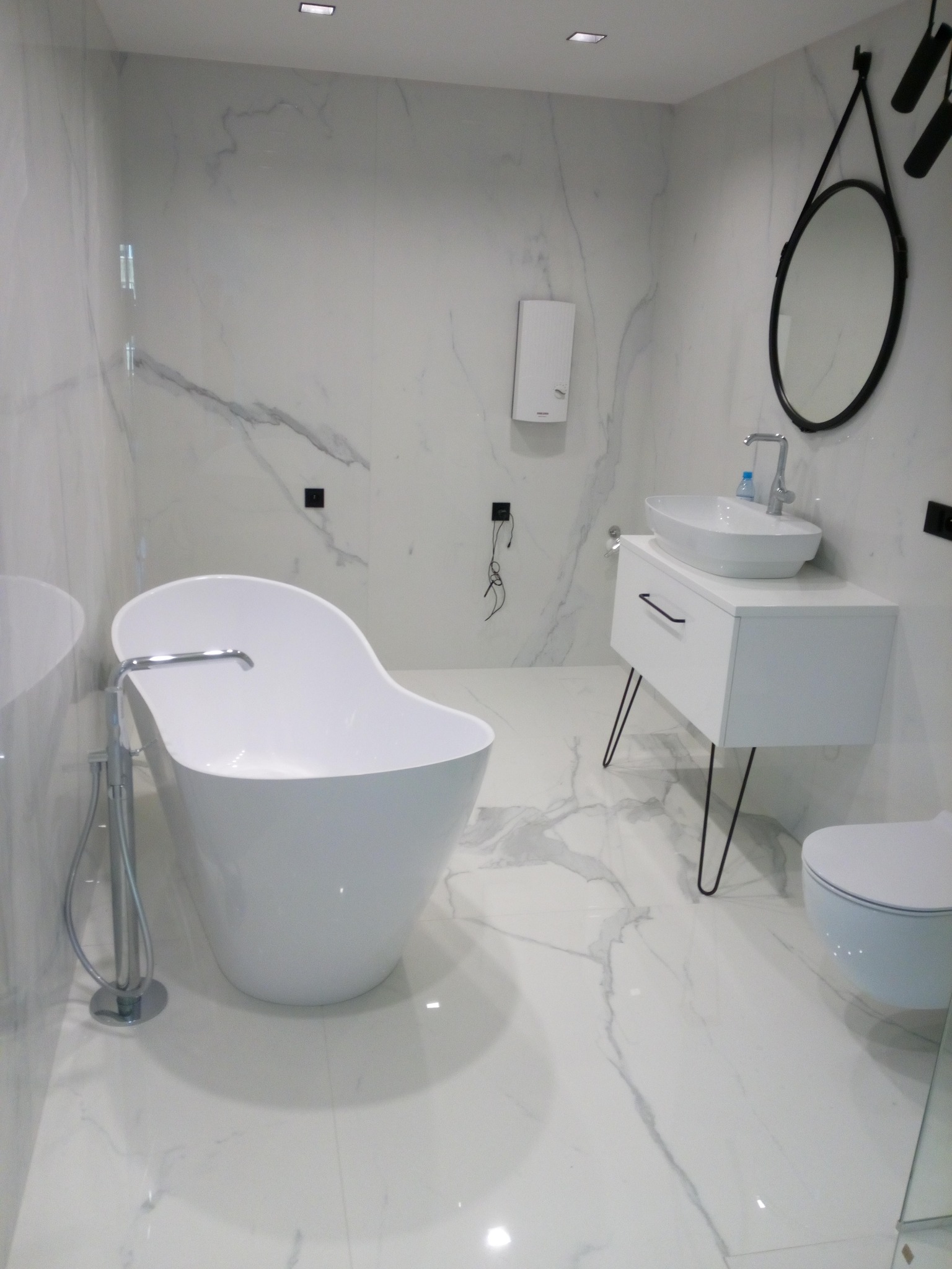 Nowoczesna łazienka z wolnostojącą wanną i umywalką na szafce, wykończona białym marmurem z szarymi żyłami, okrągłe lustro w czarnej ramie.