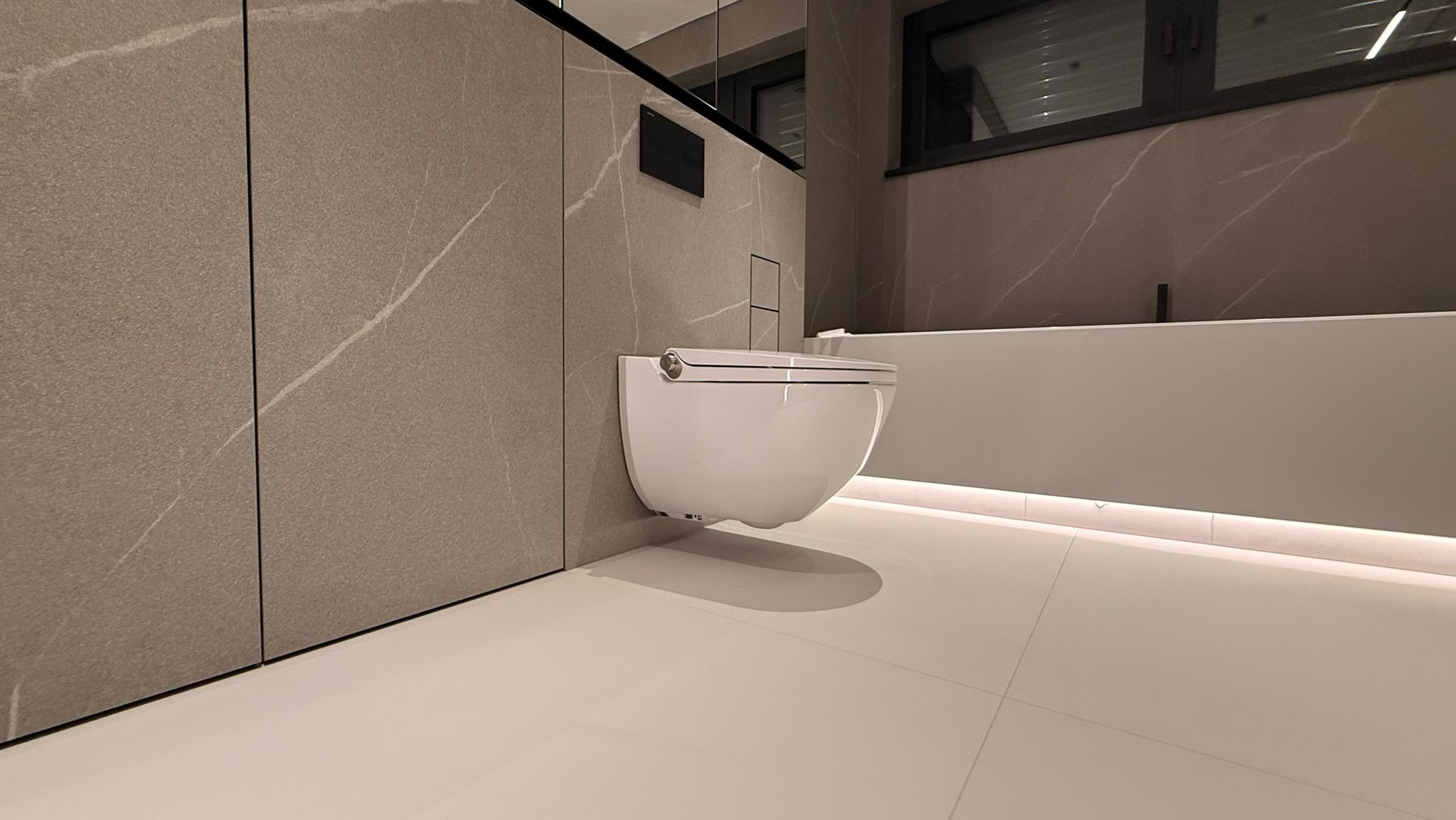 Nowoczesna toaleta podwieszana w minimalistycznej łazience z szarymi płytkami imitującymi marmur i subtelnym oświetleniem LED pod wanną. Czyste linie, elegancki design.