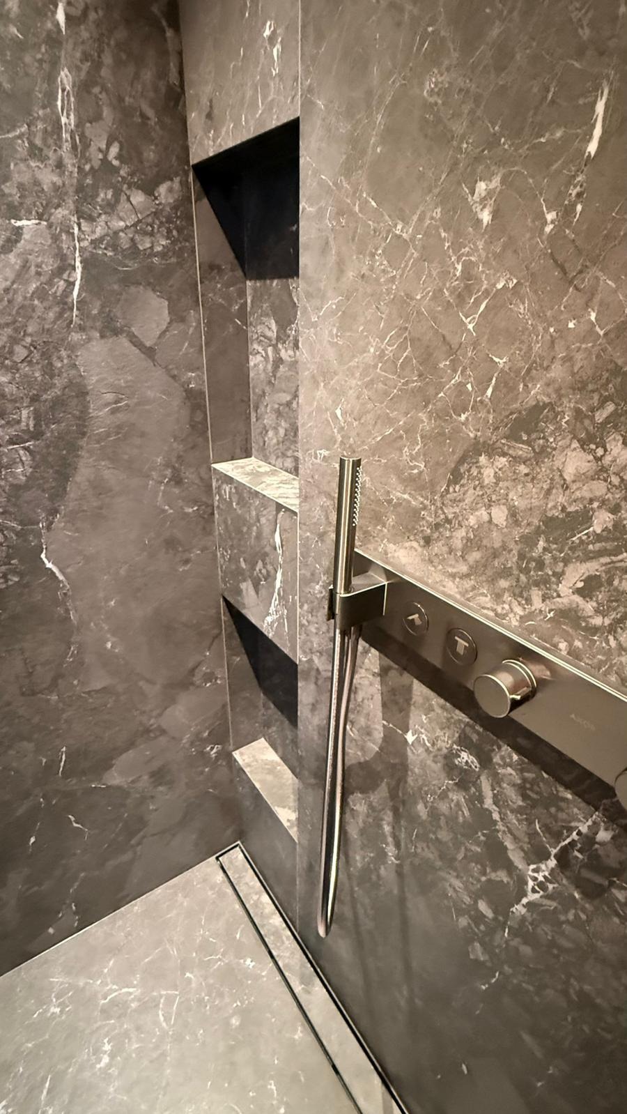 Elegancka kabina prysznicowa z szarymi płytkami imitującymi marmur, wbudowanymi półkami i nowoczesną armaturą. Minimalistyczny design z dbałością o detale.