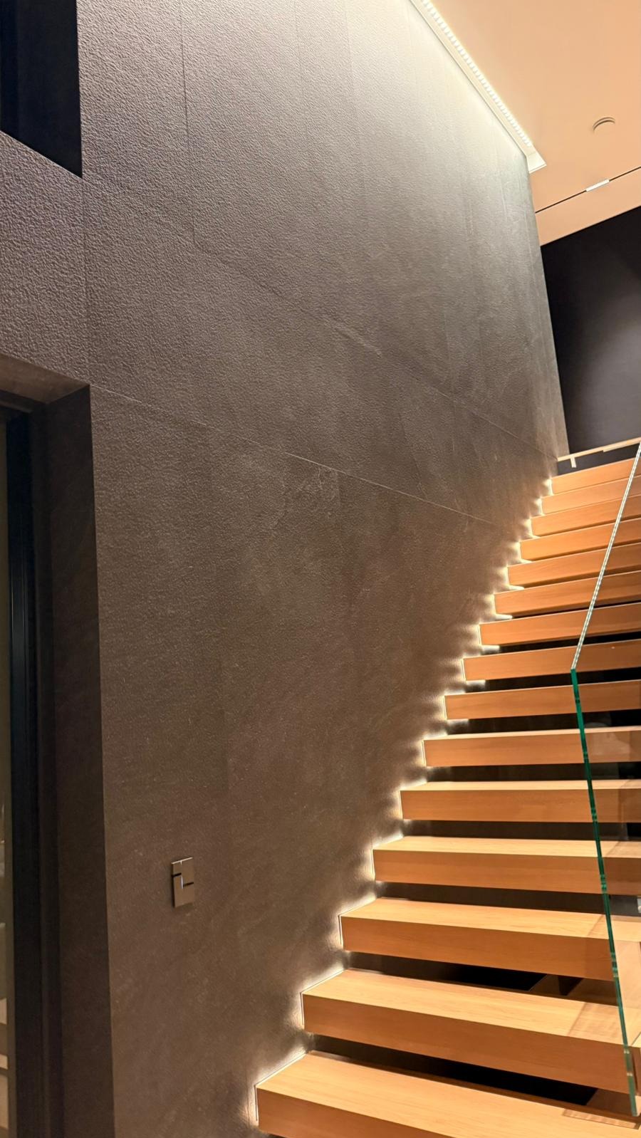 Nowoczesne schody z podświetleniem LED i szklaną balustradą, przylegające do ściany wykończonej ciemną, teksturowaną płytą. Minimalistyczny design, ciepłe oświetlenie.