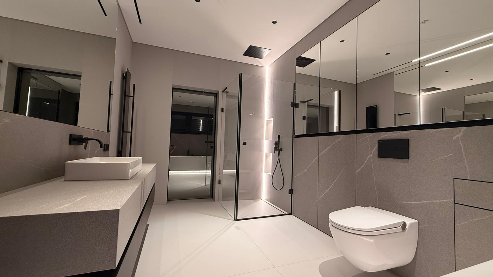 Nowoczesna łazienka w szarościach z kabiną prysznicową typu walk-in, podwieszaną toaletą i dużym lustrem. Minimalistyczny design z akcentami czerni.