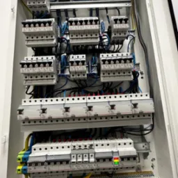 Otwarta, biała skrzynka rozdzielcza z zamontowanymi bezpiecznikami i przewodami elektrycznymi w różnych kolorach, widok z bliska na ułożone rzędy zabezpieczeń z oznaczeniami i podłączeniami.