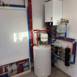 Biały piec gazowy kondensacyjny z podłączonym zasobnikiem ciepłej wody, widoczne rury instalacji grzewczej w kolorach czerwonym i niebieskim oraz rozdzielacz w szafce na ścianie.