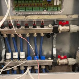 Hydro-Pro Paweł Garwoła - Rozdzielacz ogrzewania podłogowego w metalowej szafce z widocznymi zaworami, niebieskimi rurami i elektronicznym sterownikiem TECH, częściowo zamontowany w ścianie.