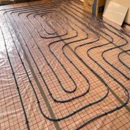 Niebieskie rury ogrzewania podłogowego rozłożone na folii z czerwoną kratką, mocowane czarnymi uchwytami, widoczny karton po systemie grzewczym w tle, w pomieszczeniu w stanie surowym.