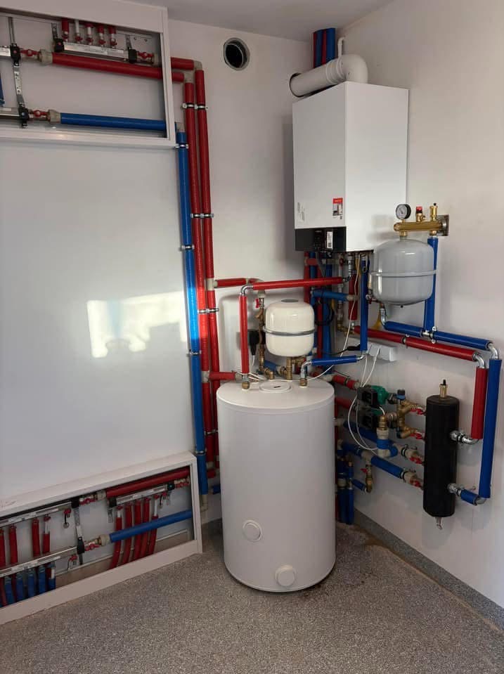 Biały piec gazowy kondensacyjny z podłączonym zasobnikiem ciepłej wody, widoczne rury instalacji grzewczej w kolorach czerwonym i niebieskim oraz rozdzielacz w szafce na ścianie.