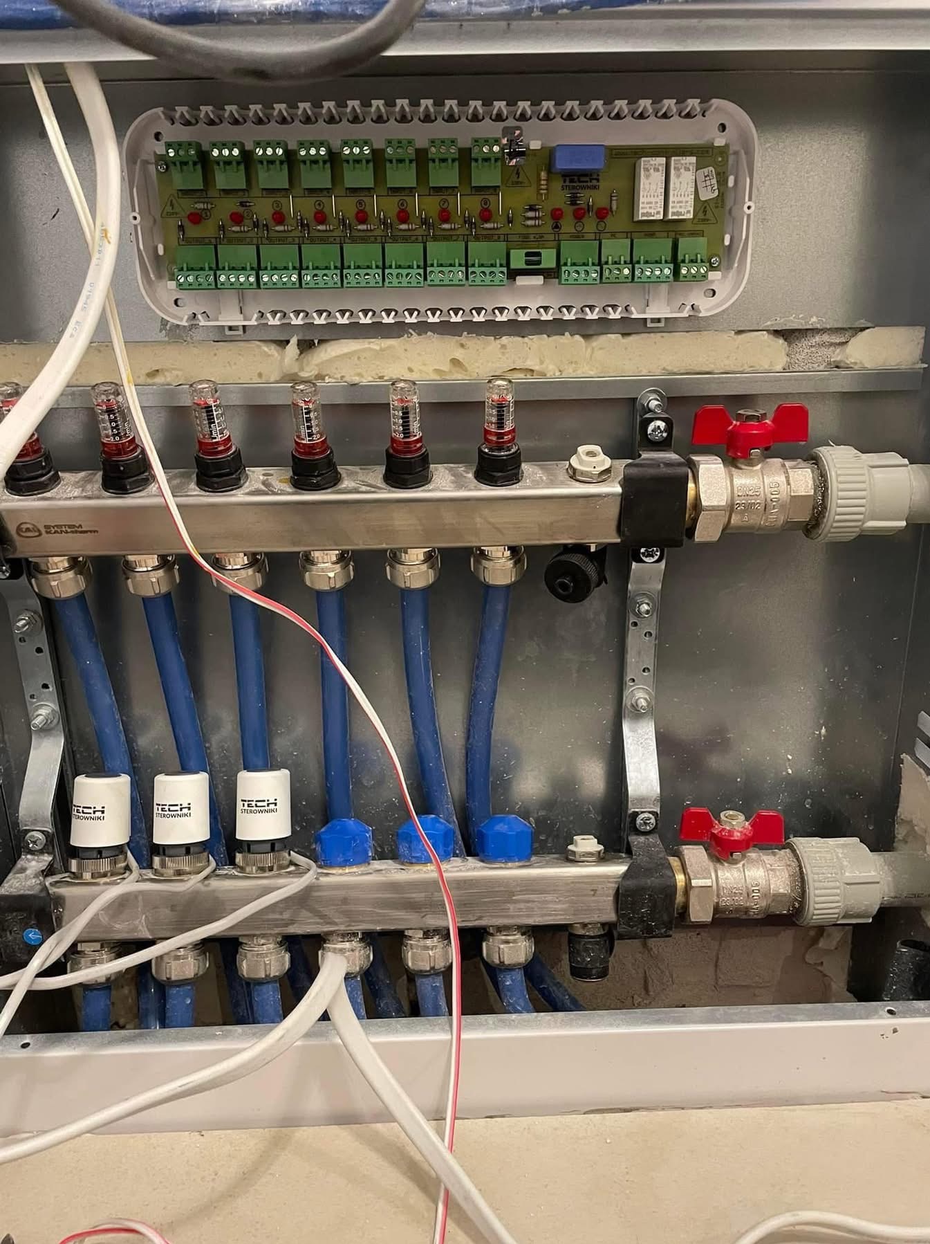 Rozdzielacz ogrzewania podłogowego w metalowej szafce z widocznymi zaworami, niebieskimi rurami i elektronicznym sterownikiem TECH, częściowo zamontowany w ścianie.
