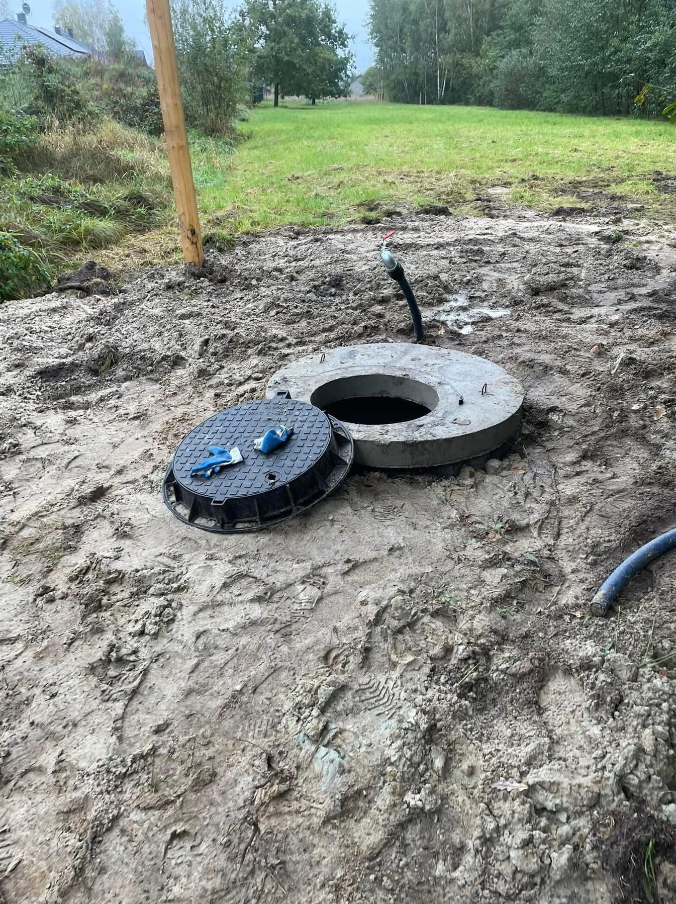 Otwarta studzienka kanalizacyjna z betonowym kręgiem i leżącą obok pokrywą, na której znajdują się niebieskie rękawice robocze, widoczne ślady w błocie po pracach ziemnych, w tle trawa i drzewa.