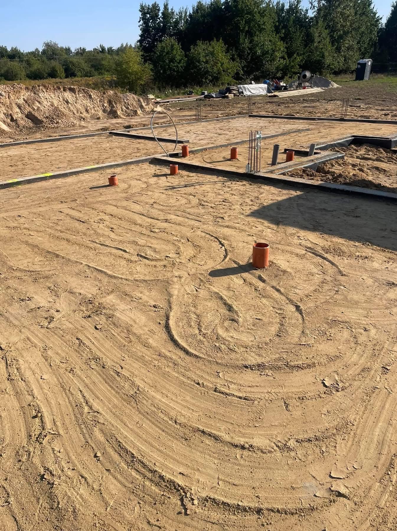 Przygotowanie podłoża pod budowę domu: widoczne fundamenty, wystające rury kanalizacyjne i ślady po ułożeniu instalacji w piasku.