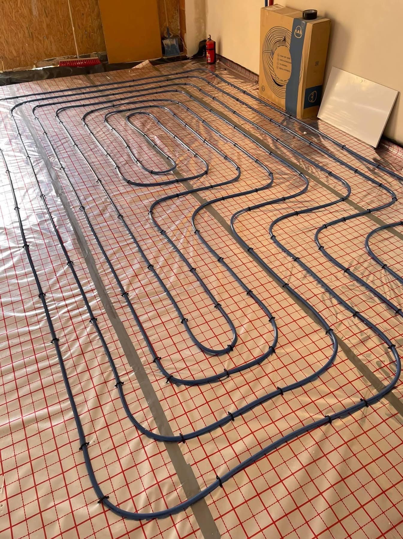 Niebieskie rury ogrzewania podłogowego rozłożone na folii z czerwoną kratką, mocowane czarnymi uchwytami, widoczny karton po systemie grzewczym w tle, w pomieszczeniu w stanie surowym.