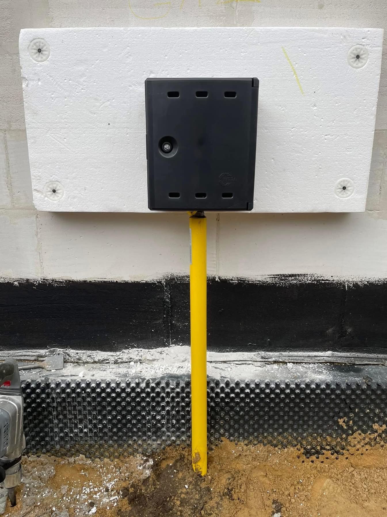 Żółta rura gazowa wchodząca do czarnej skrzynki na ścianie budynku, widoczna warstwa izolacji i folia kubełkowa przy gruncie, w tle ściana z białym styropianem.