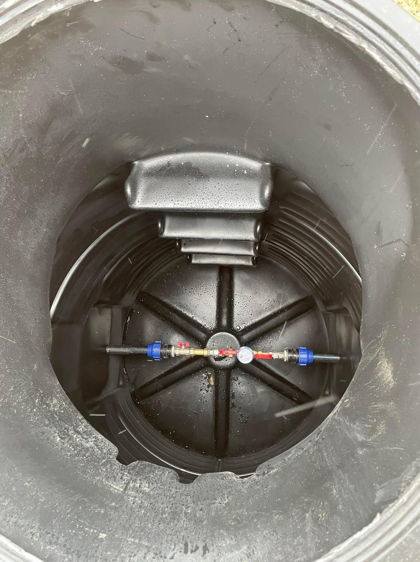 Wnętrze czarnego zbiornika z widocznymi przyłączami wodnymi, zaworami i manometrem, z lekkimi śladami wilgoci na elementach.