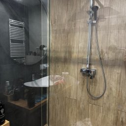 F.U BLASK CZYSTOŚCI PAULINA CHĄDZYŃSKA - Szklana kabina prysznicowa z deszczownicą i baterią termostatyczną na drewnopodobnej ścianie, widoczna umywalka i grzejnik drabinkowy w tle.