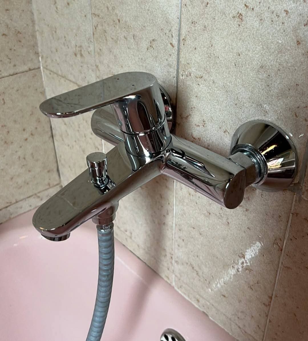 Bateria wannowa chromowana, zamontowana na płytkach ceramicznych beżowego koloru, z widocznym wężem prysznicowym i fragmentem różowej wanny.