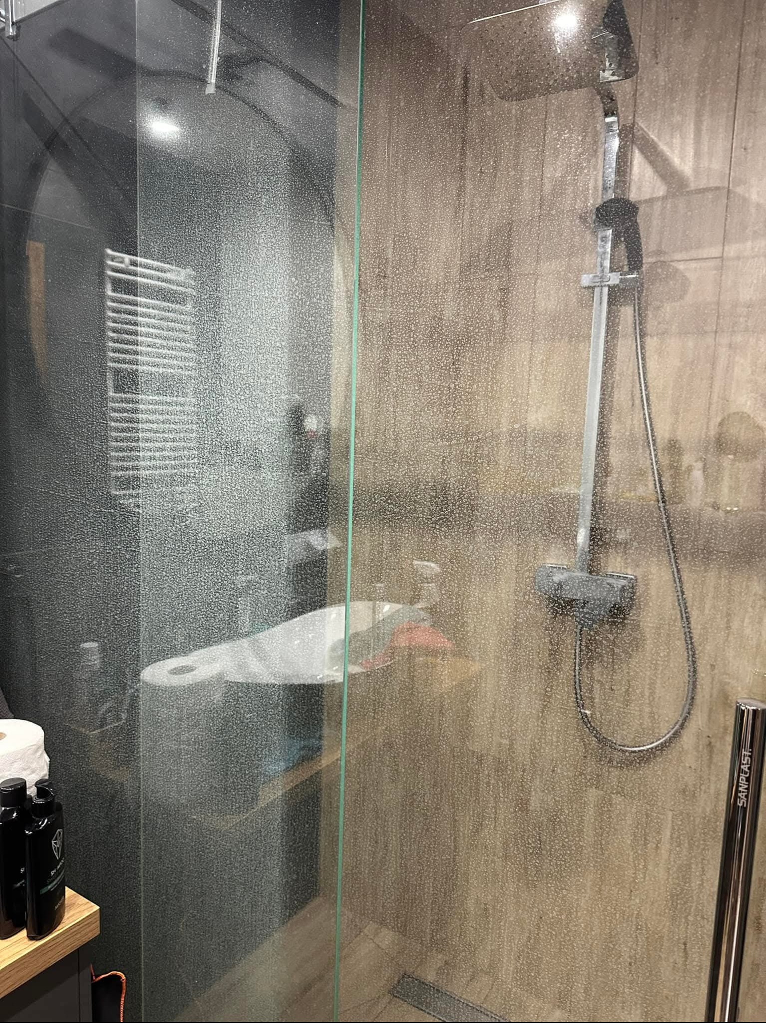Kabina prysznicowa ze śladami po wodzie na szklanych drzwiach, widoczna słuchawka prysznicowa i półka z kosmetykami w tle.
