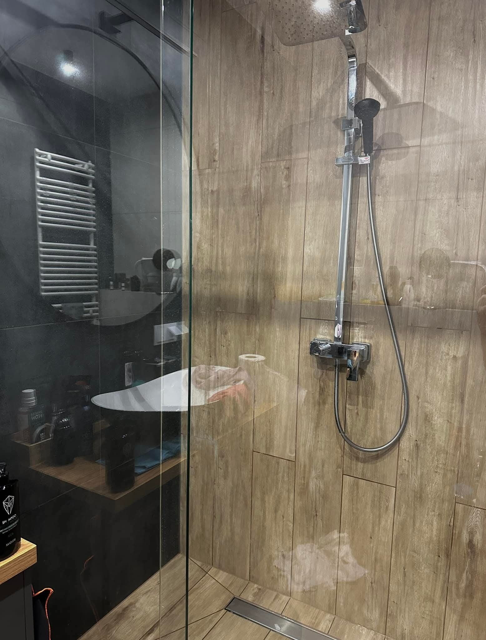Szklana kabina prysznicowa z deszczownicą i baterią termostatyczną na drewnopodobnej ścianie, widoczna umywalka i grzejnik drabinkowy w tle.