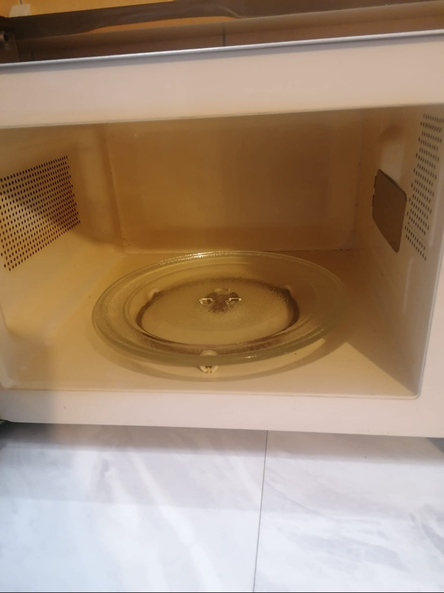 Wnętrze beżowej kuchenki mikrofalowej z okrągłym, szklanym talerzem obrotowym; widoczne perforowane ściany boczne i plamy zabrudzeń.