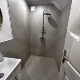 Nowoczesna kabina prysznicowa z szarymi płytkami imitującymi kamień, odpływ liniowy, biała umywalka z szafką i wiszącym ręcznikiem, oświetlenie punktowe na suficie skośnym.