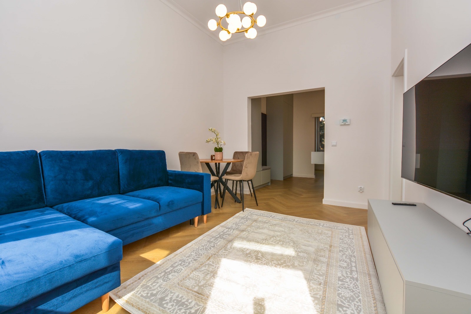 Salon z niebieską sofą narożną, wzorzystym dywanem, okrągłym stołem z krzesłami i nowoczesną lampą sufitową, parkiet w jodełkę.