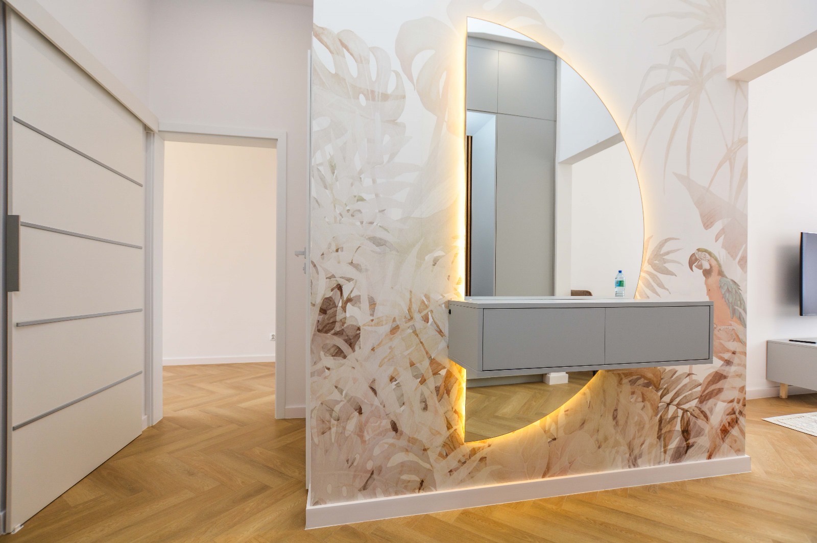 Nowoczesny przedpokój z lustrem w kształcie koła podświetlonym LED, szafką wiszącą, tapetą w motywy tropikalne z papugą i parkietem w jodełkę.