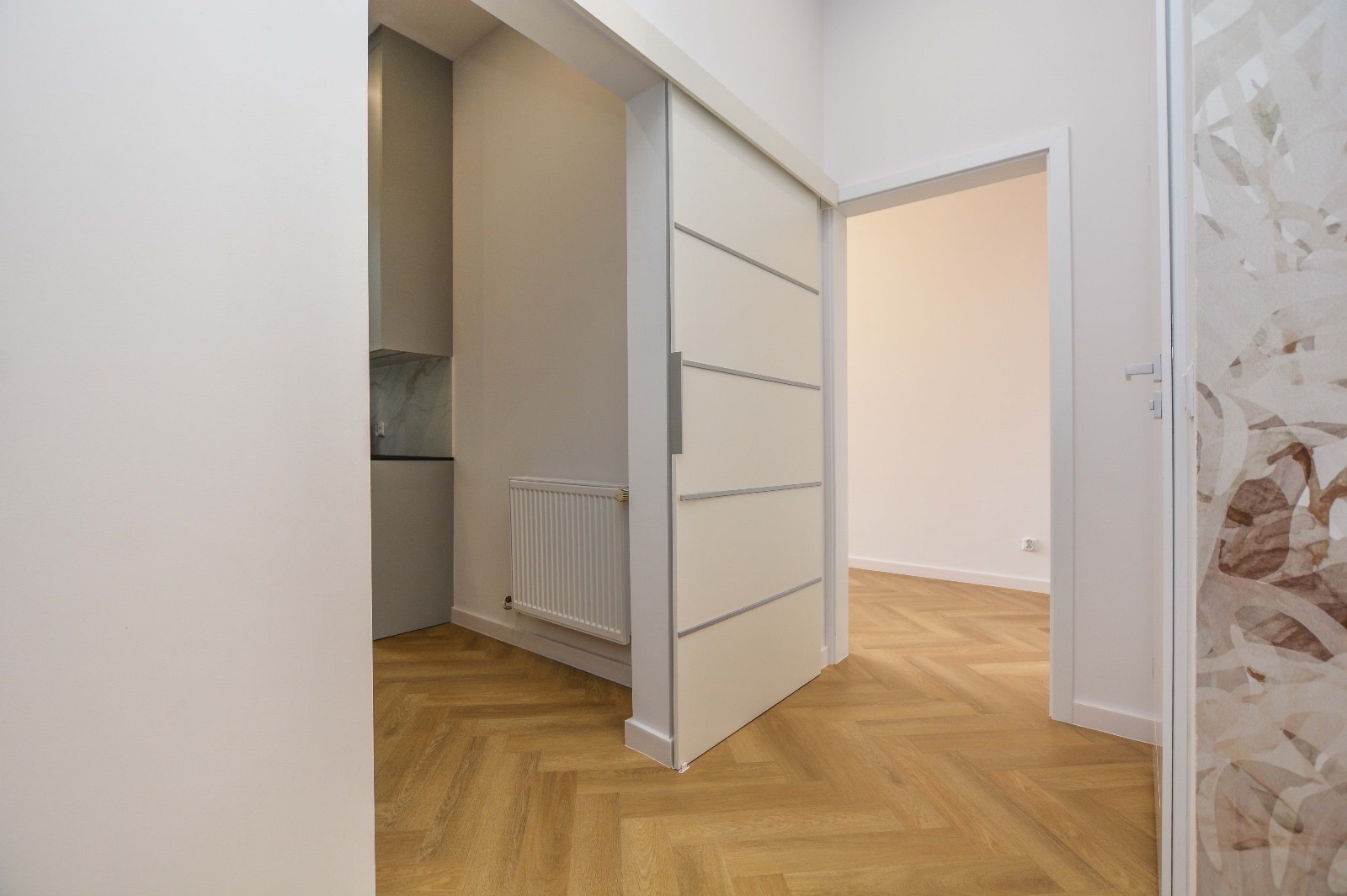 Nowoczesne wnętrze mieszkania z parkietem w jodełkę, częściowo otwarte przesuwne drzwi z czterema poziomymi listwami i dekoracyjna folia na szklanych drzwiach po prawej.