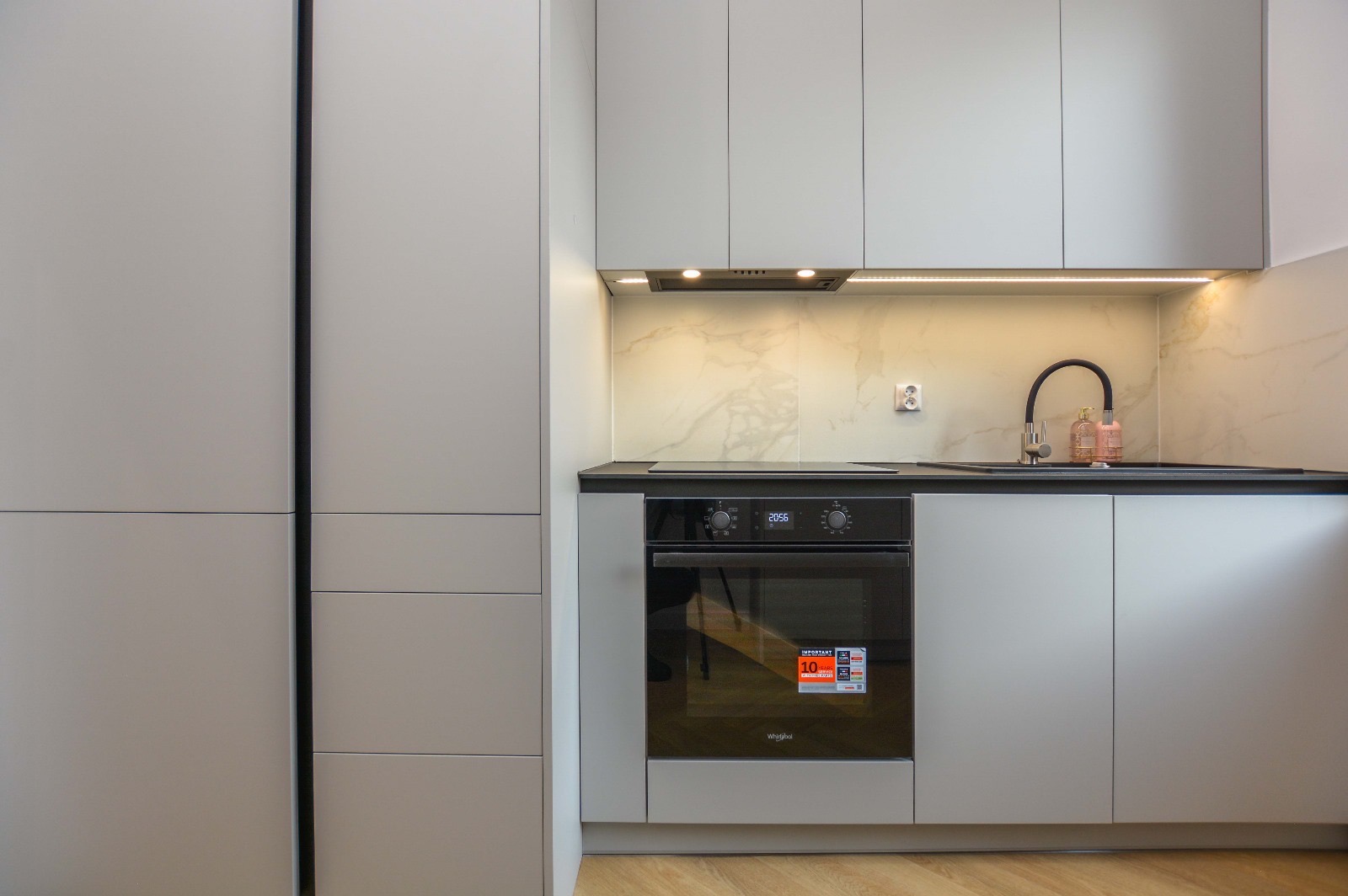 Nowoczesna kuchnia w odcieniach szarości z zabudową meblową, piekarnikiem z naklejką promocyjną, czarnym blatem i marmurowym backsplashiem podświetlonym ledami.