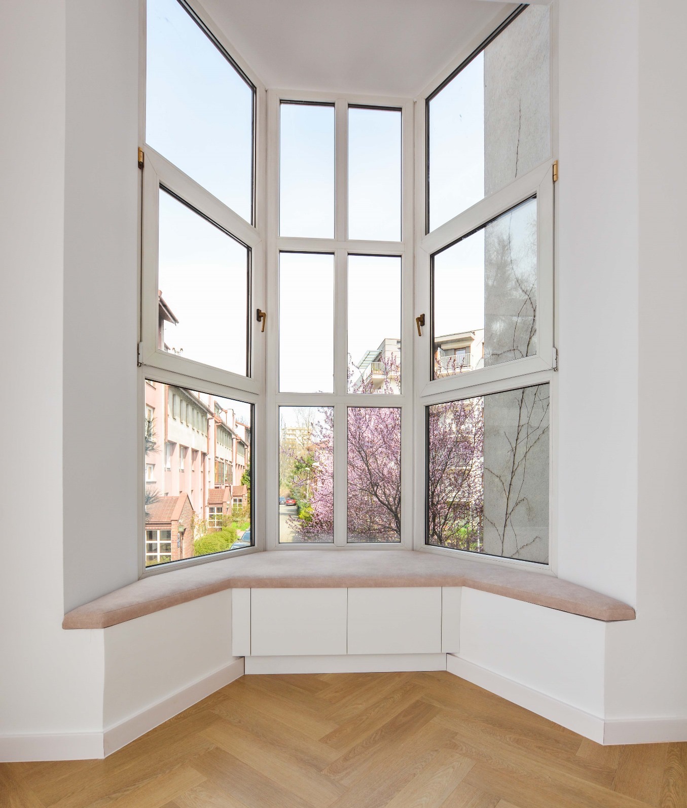 Jasne wnętrze z wykuszem okiennym, drewnianą podłogą w jodełkę i widokiem na kwitnące drzewa przez otwarte okna.