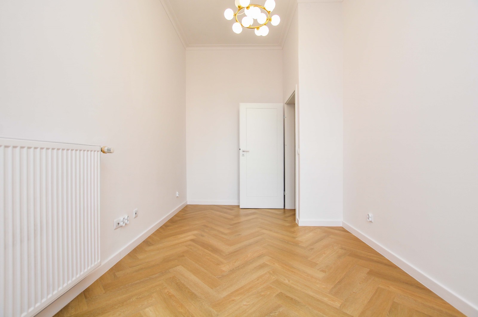 Puste, odnowione wnętrze z parkietem ułożonym w jodełkę, białymi ścianami, grzejnikiem i nowoczesną lampą sufitową w stylu retro, widok na otwarte białe drzwi.