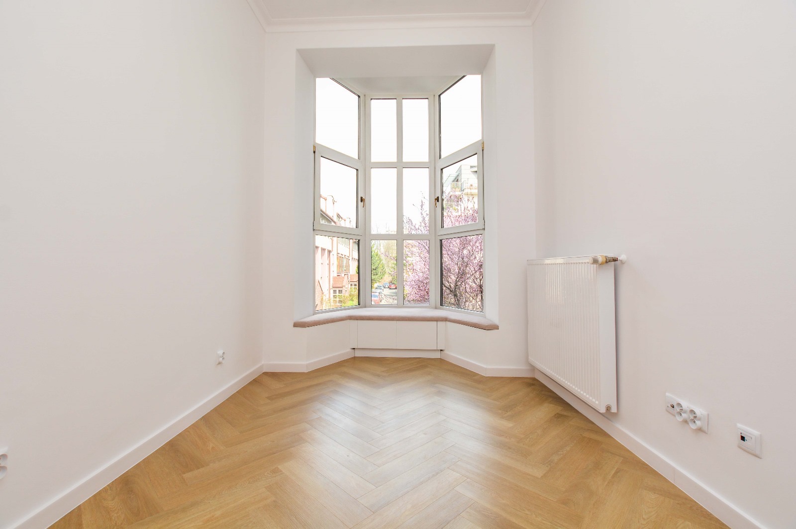 Pusty pokój z parkietem w jodełkę, białymi ścianami i wykuszem z widokiem na kwitnące drzewa.