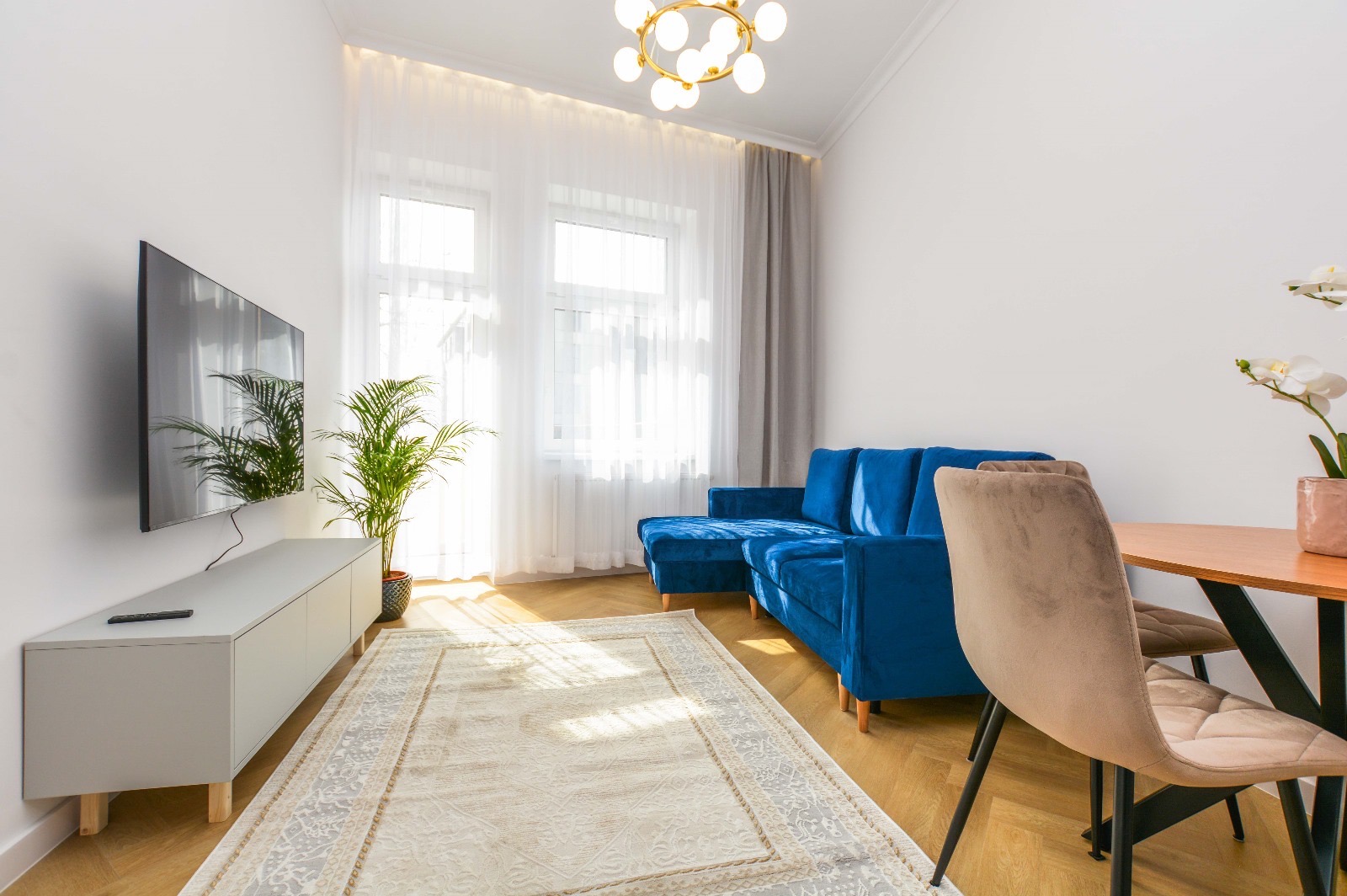 Jasne wnętrze salonu z parkietem ułożonym w jodełkę, niebieską sofą z szezlongiem, szarym stolikiem RTV z telewizorem, kremowym dywanem i okrągłym stołem z krzesłami w odcieniach beżu.