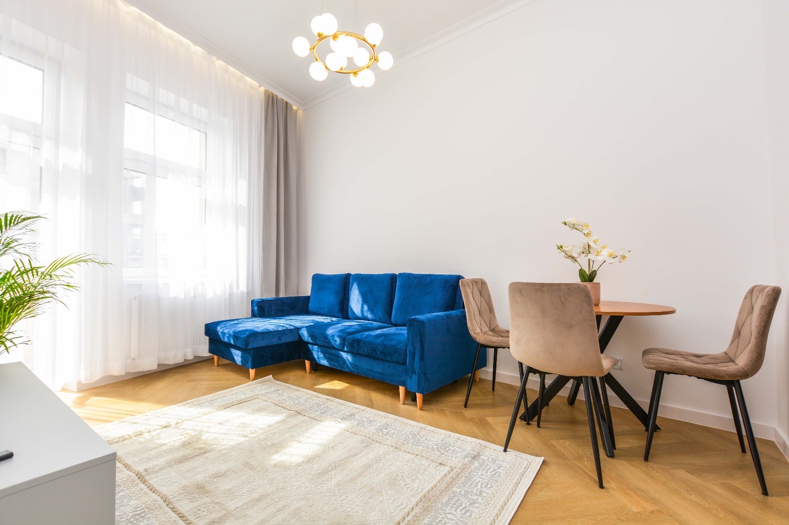 Jasne wnętrze pokoju dziennego z niebieską sofą narożną, okrągłym stołem z krzesłami i wzorzystym dywanem na parkiecie w jodełkę, oświetlone nowoczesną lampą sufitową.