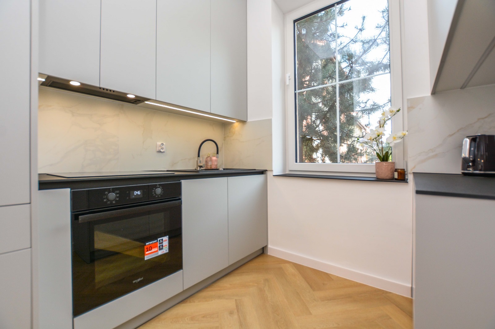 Nowoczesna, jasna kuchnia z szarymi szafkami, czarnym blatem i piekarnikiem Whirlpool z naklejką informacyjną, wykończona marmurowymi płytkami i parkietem w jodełkę, z oknem z widokiem na drzewo...