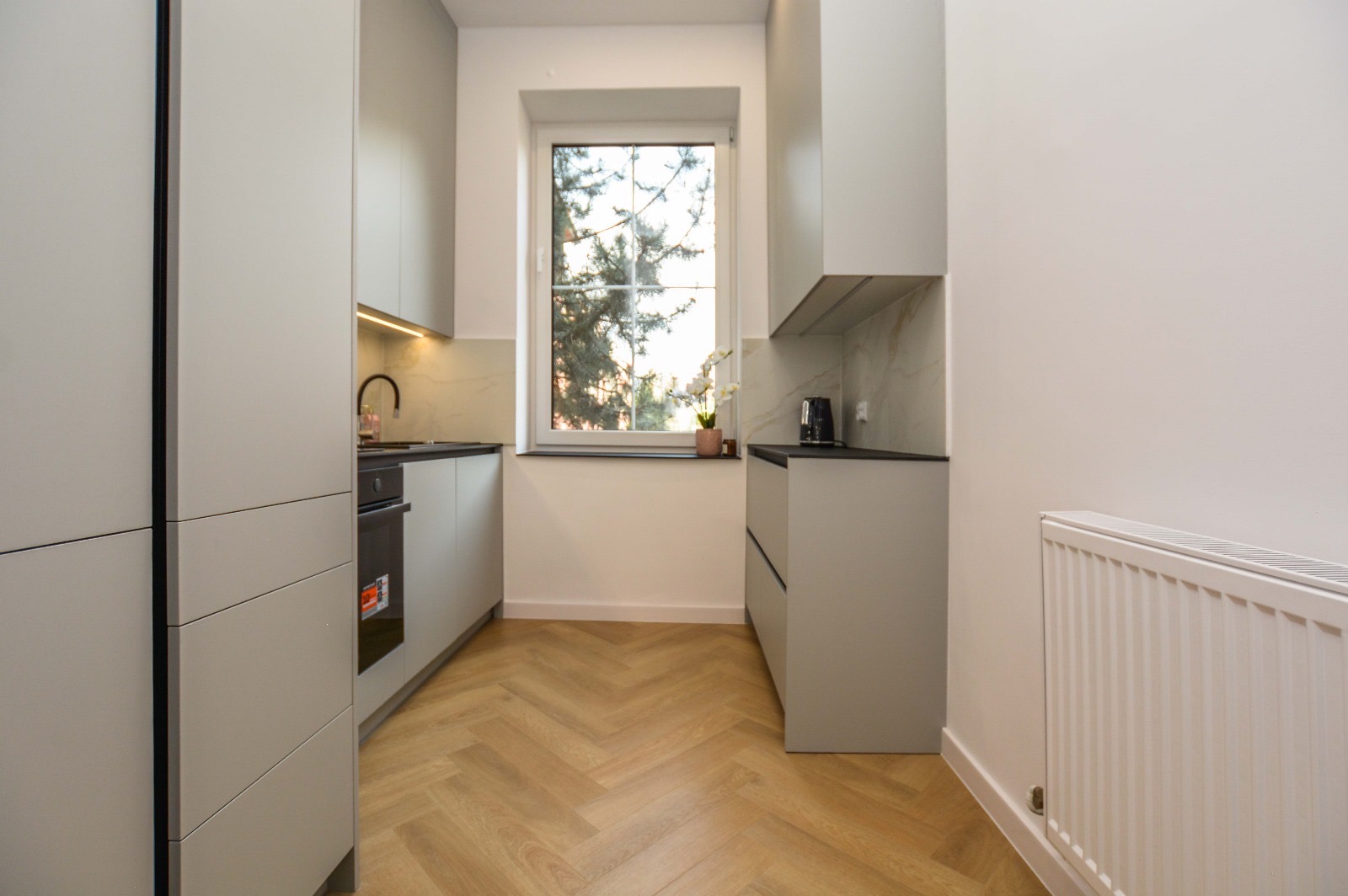 Nowoczesna kuchnia w odcieniach szarości z drewnianą podłogą ułożoną w jodełkę, oświetleniem LED pod szafkami i białym grzejnikiem na ścianie.