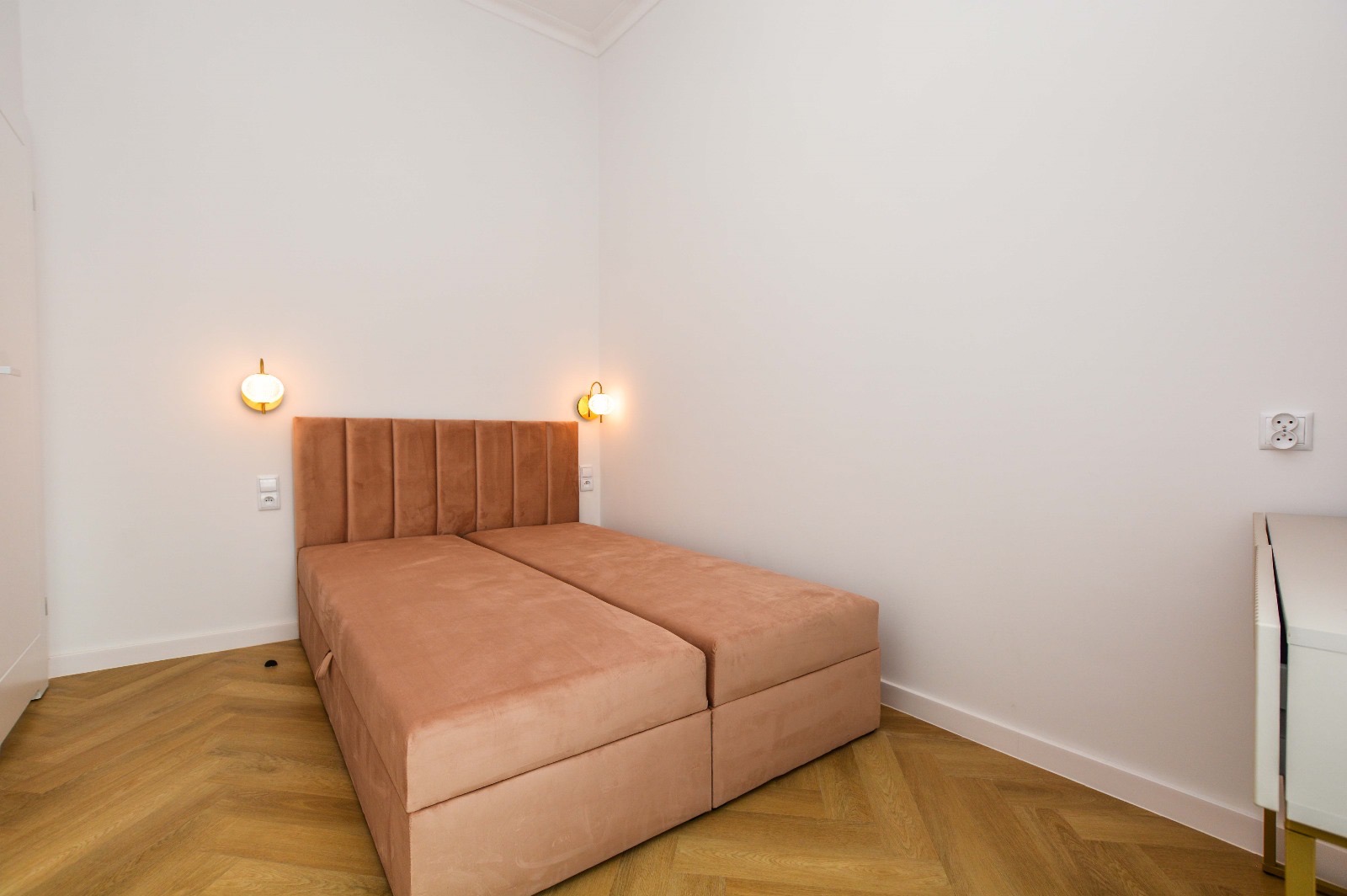 Dwa pojedyncze łóżka z brązowym, welurowym wezgłowiem i pojemnikami, ustawione obok siebie na parkiecie ułożonym w jodełkę, oświetlone przez dwie złote lampki ścienne, w minimalistycznym wnętrzu...