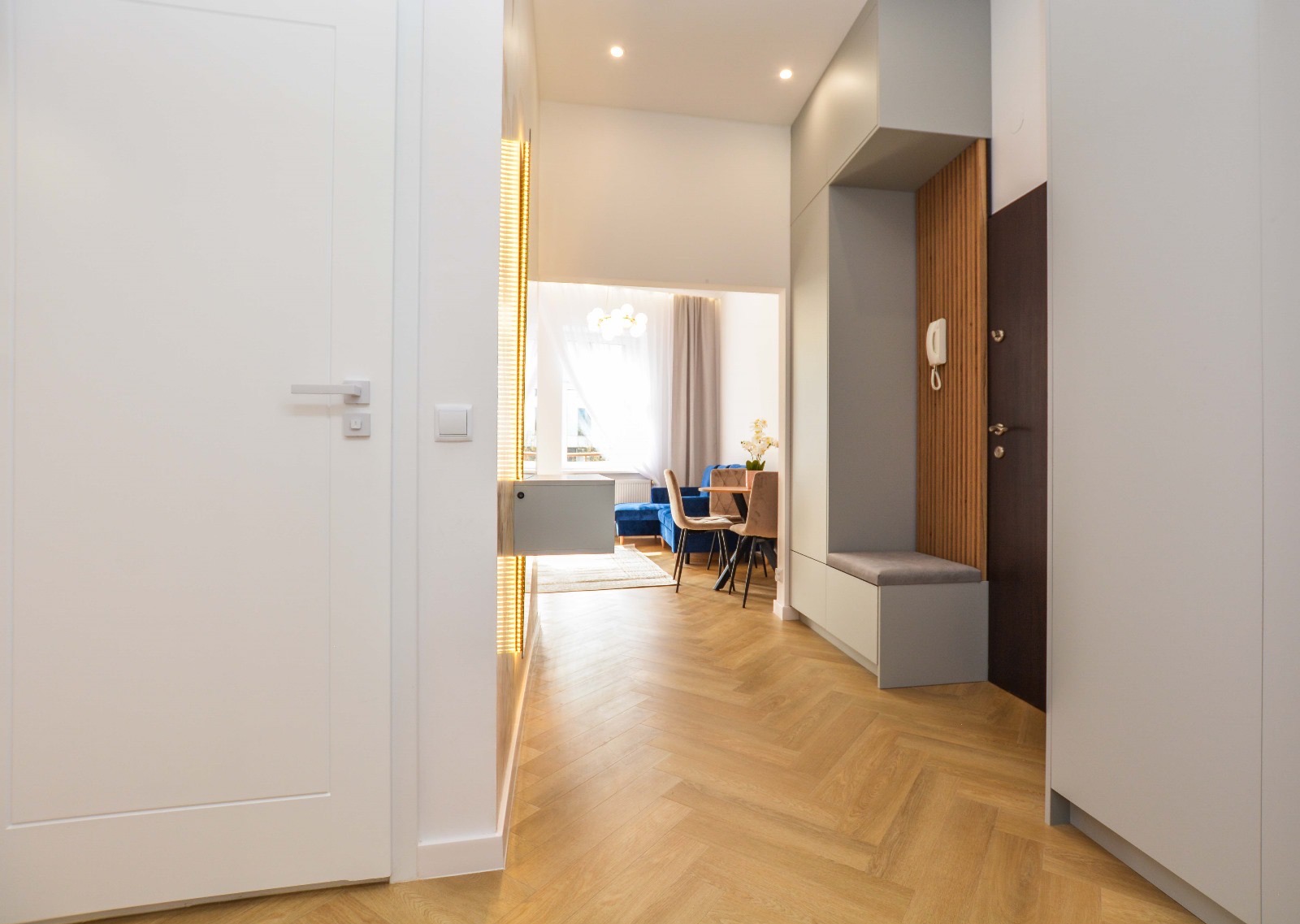 Jasne wnętrze mieszkania z parkietem w jodełkę, widok na salon z niebieską sofą i jadalnię, w przedpokoju zabudowa z siedziskiem i wieszakiem na ścianie z drewnianymi panelami.