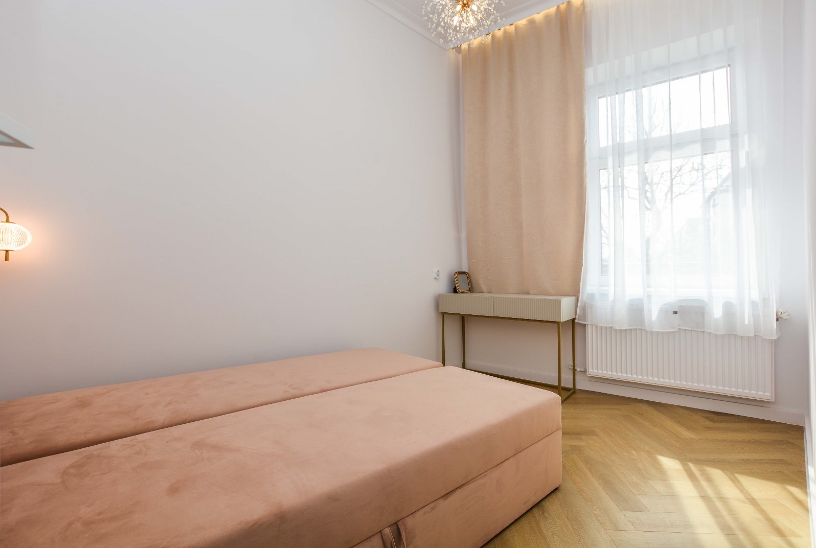 Jasne wnętrze sypialni z dwoma tapczanami w kolorze pudrowego różu, parkietem w jodełkę, konsolą z ryflowanymi frontami i złoconymi nogami, oraz okrągłą lampą na ścianie.