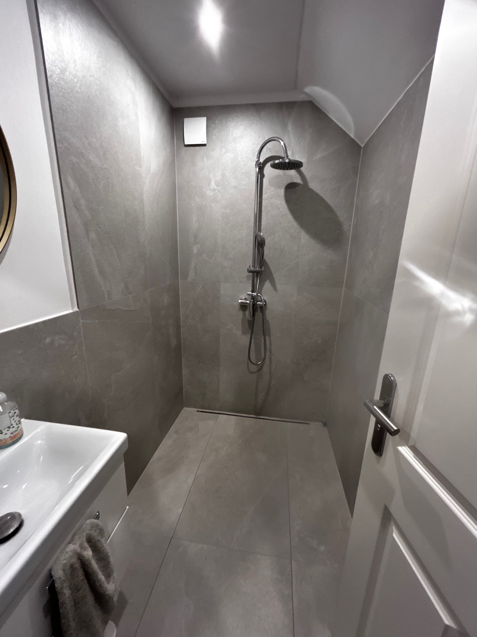 Nowoczesna kabina prysznicowa z szarymi płytkami imitującymi kamień, odpływ liniowy, biała umywalka z szafką i wiszącym ręcznikiem, oświetlenie punktowe na suficie skośnym.