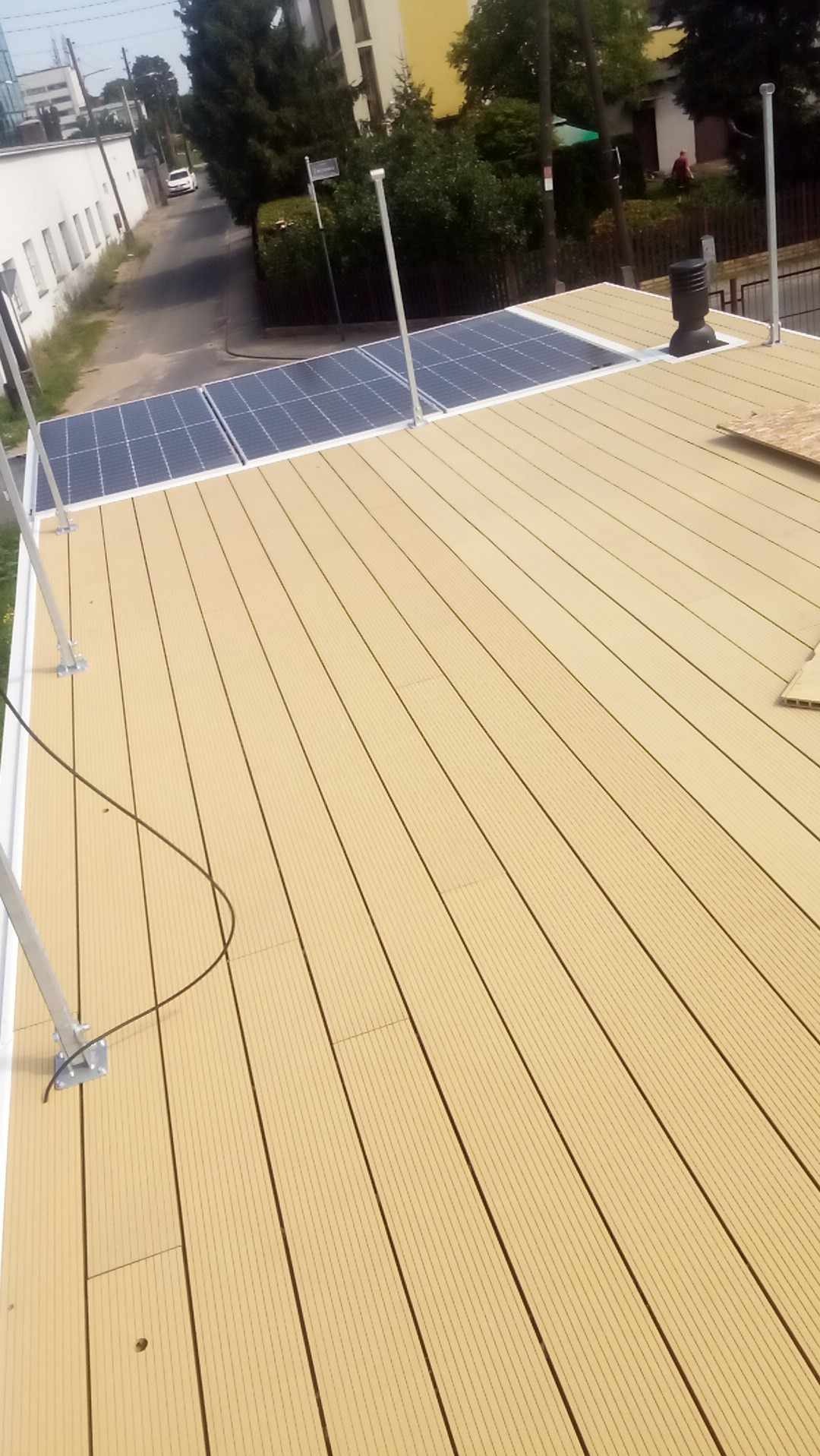 Beżowa deska tarasowa z panelami słonecznymi na dachu budynku, widok z góry. Elementy montażowe widoczne. Ulica i budynki w tle.