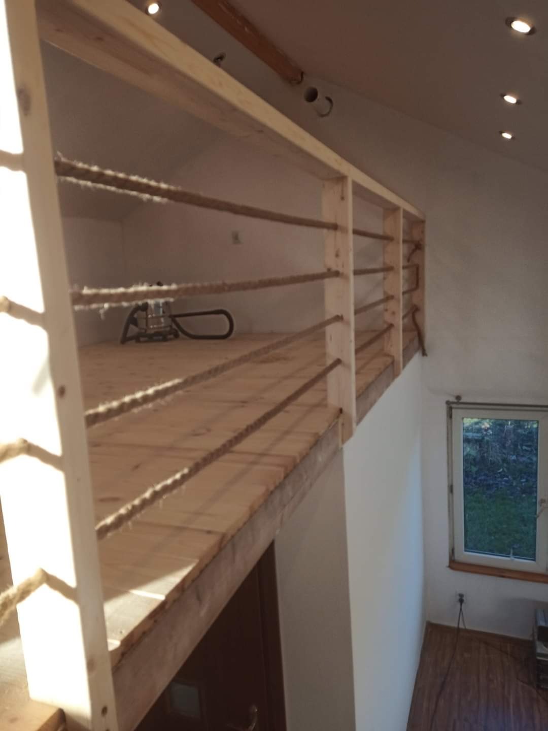 Drewniana antresola z linowymi barierkami w jasnym wnętrzu poddasza, widok z dołu, okno w tle. Surowy styl wykończenia, widoczne niedoskonałości drewna.