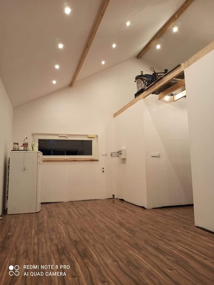 Jasne wnętrze ze skośnym sufitem, drewnianymi belkami i nową podłogą z paneli. Widoczne oświetlenie punktowe i okno. Minimalistyczny wystrój, białe ściany.