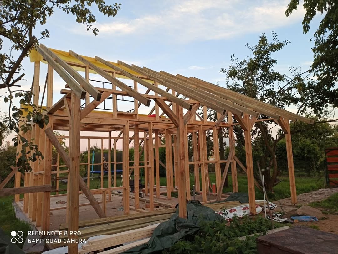 Drewniana konstrukcja szkieletowa budynku w trakcie budowy, widok z zewnątrz, z elementami żółtej membrany dachowej i zielonym otoczeniem.