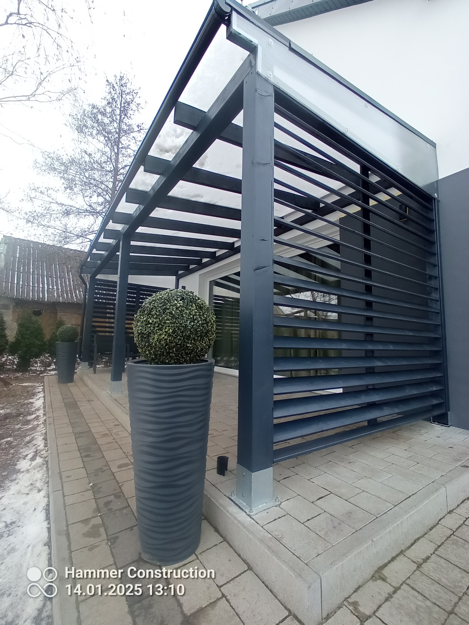 Nowoczesna, szara pergola z żaluzjami i donicami z bukszpanem, na tle kostki brukowej i budynku. Minimalistyczny design, zimowa sceneria z resztkami śniegu.